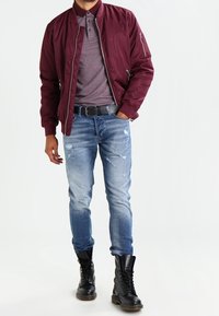 Bordowa kurtka typu bomber, teksturowane szare polo, jasnoniebieskie podniszczone dżinsy oraz czarne bojówki ze sznurowadłami i metalicznymi dodatkami.