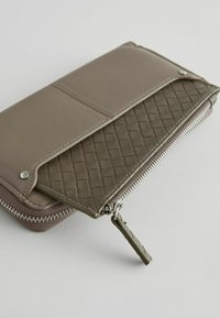 Rektangulær pung i dæmpet taupe læder med et vævet indre. Sølvlynlåser og -hardware, med en lille tekstureret lynlåspull.