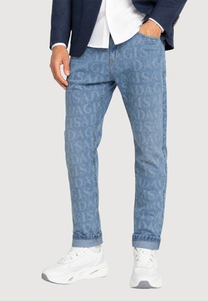 Uomo che indossa jeans azzurri chiari con motivo a testo su tutta la superficie, blazer blu navy, camicia bianca e sneakers bianche, in piedi con una mano in tasca.