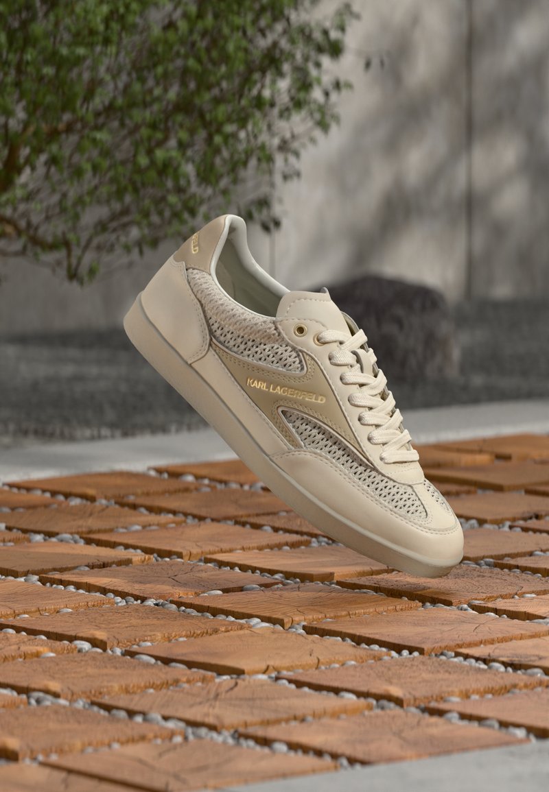 Μπεζ sneaker Karl Lagerfeld με διχτυωτές και δερμάτινες λεπτομέρειες, κορδόνια και χρυσό λογότυπο, τοποθετημένο στον αέρα πάνω από ξύλινα πλακάκια σε εξωτερικό χώρο.
