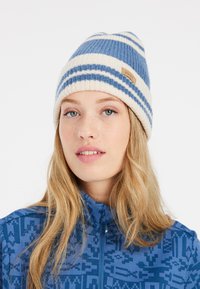 Bonnet tricoté avec un motif rayé bleu et crème. Mélange de laine doux avec une finition texturée. Présente une petite étiquette beige sur le côté.