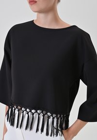 Top corto nero realizzato in tessuto liscio, con scollatura ampia e dettagli di frange a nappine sull'orlo. Accenti in nero intrecciato.