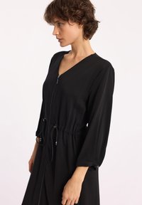 Czarna sukienka z zamkiem z przodu, dekoltem w kształcie litery V, ze zmarszczonym wcięciem w talii i rękawami o długości trzech ćwierć. Posiada miękką, teksturowaną tkaninę oraz sznurki do regulacji.