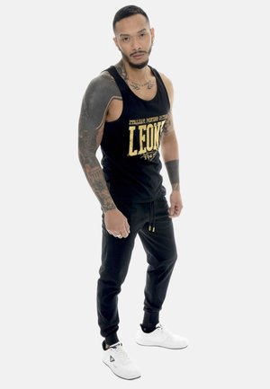 Homme avec des tatouages portant un débardeur noir avec des inscriptions dorées, un pantalon de jogging noir et des baskets blanches, debout sur fond blanc.