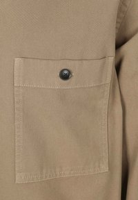 Beige stof met een genuanceerde weving, voorzien van een borstzak die met een zwarte knoop is bevestigd. Eenvoudig ontwerp met zichtbare stikseldetails.