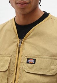 Närbild av en person som bär en beige zip-up jacka med bröstfickor och en Dickies logotyp-lapp på höger ficka.