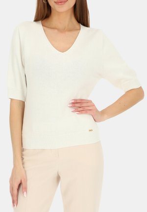 Kobieta ubrana w biały sweter z krótkim rękawem i dekoltem w kształcie litery V oraz beżowe spodnie, pozująca z jedną ręką na biodrze, a drugą wzdłuż ciała.