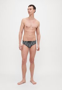 Chiloți de înot negri cu un model de text alb, design ajustat, textură netedă și curea elastică, purtați de un model masculin în picioare.