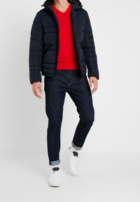 Giacca imbottita blu navy con cappuccio, maglione a scollo a V rosso sotto, jeans scuri e sneakers bianche. Presenta una chiusura con cerniera e tasche laterali.