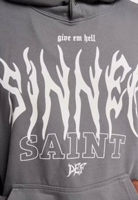 Grå hettegenser med hvit, flammeaktig tekst som leser "SINNER" og "SAINT," med "give em hell" over og en liten logo under.