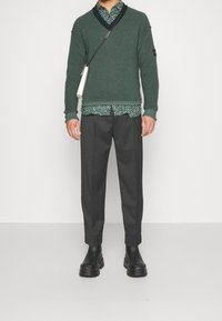 Pull vert en maille, col en V noir, chemise à motif floral, pantalon gris et bottines noires. Un sac bandoulière complète le look.