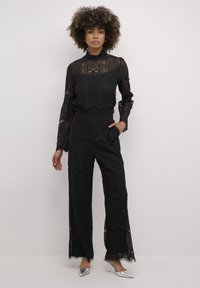Juodas nėriniuotas jumpsuit'as su ilgomis rankovėmis, sudėtingais raštais ir plačiomis kelnėmis su banguotu kraštu. Sidabrinės aukštakulnės bateliai užbaigia įvaizdį.