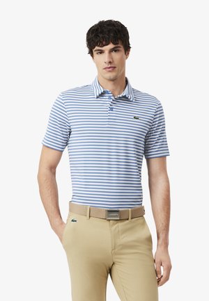 Uomo che indossa una polo a maniche corte a righe blu e bianche e pantaloni beige con cintura beige, in piedi con le mani in tasca.