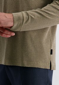 Texturiertes Langarmshirt in einem khaki- und cremefarbenen Muster, mit Seitenschlitzen und einem Etikett. Der Stoff wirkt gewebt und strapazierfähig.