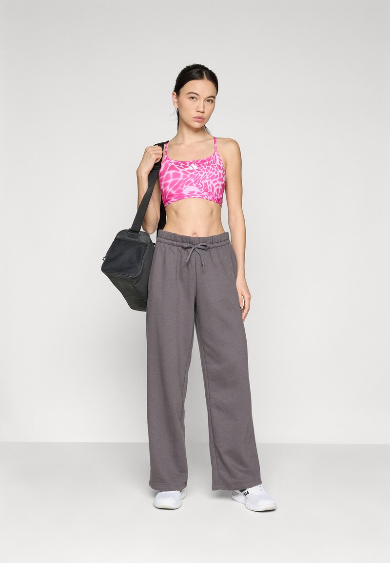 Rosa gemusterter Sport-BH, graue locker sitzende Sweatpants mit Kordelzug und weiße Sportschuhe; Modell hält eine schwarze Tasche.