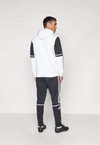 White and black tracksuit with a hood, featuring contrasting sleeves, three white stripes on pants, and black athletic shoes.