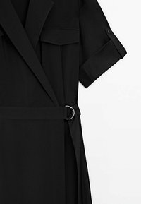 Schwarzes Kleid mit Gürtel, aufgerollten Kurzärmeln, eingeschnittenem Kragen und Klapptaschen auf der Brust, vor weißem Hintergrund.