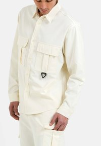 Camisa de manga larga color crema con dos bolsillos frontales, cuello abotonado y detalle triangular de hardware negro. Tejido texturizado.