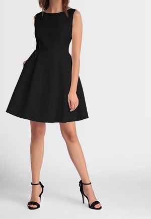 Kobieta w czarnej sukience bez rękawów o kroju litery A z kieszeniami, ubrana w czarne sandały na wysokim obcasie z paskiem wokół kostki, stojąca na tle jednolitego tła.