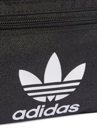 Gros plan sur un tissu noir avec le logo trèfle blanc d'Adidas et une fermeture éclair au-dessus, sur une surface texturée.