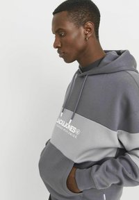 Grau farblich abgesetzter Hoodie mit einer großen frontalen Tasche und verstellbarem Zugband. Mit weißem Logo-Text auf der Brust und weichem Stoffgefühl.