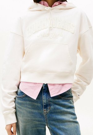 Person trägt einen cremefarbenen, kurz geschnittenen Hoodie mit der Aufschrift "Tommy Jeans New York", darüber ein rosa Hemd und blaue Jeans.