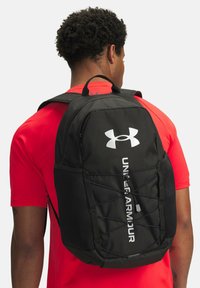 Juodas kuprinės modelis su "Under Armour" logotipu, turintis tinklines šonines kišenes, reguliuojamus dirželius ir elastingą virvę priekyje.