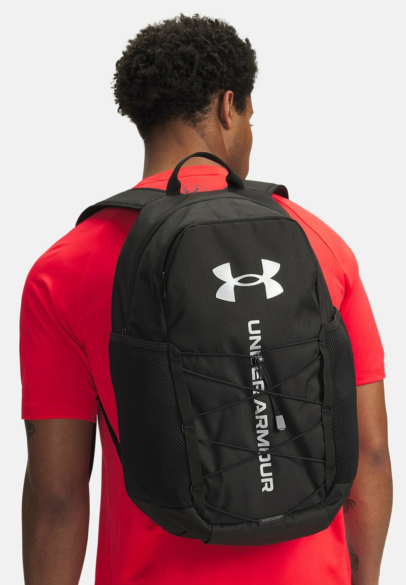 Juodas kuprinės modelis su "Under Armour" logotipu, turintis tinklines šonines kišenes, reguliuojamus dirželius ir elastingą virvę priekyje.
