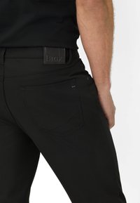 Zwarte broek van gladde stof, met een standaard achterzak en een opvallende zwarte band met het merk "Brax" in reliëf.