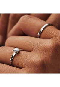 Silberring mit einem quadratischen Diamantmittelstück, glatte Oberfläche und gebogene Band, daneben ein strukturiertes, gemustertes Silberband an einer Hand.