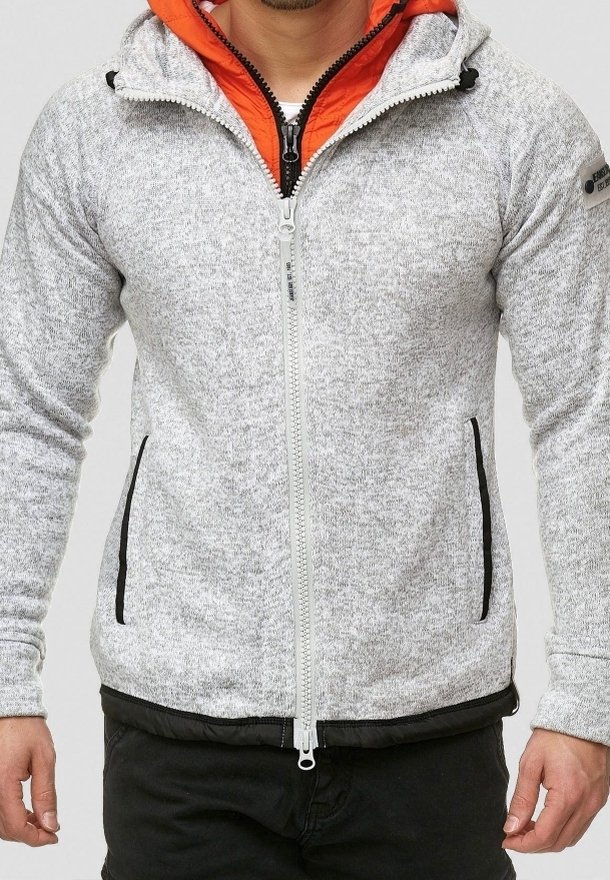 Homme portant un sweat à capuche zippé gris clair avec une fermeture éclair noire et des bordures de poche noires, une couche intérieure orange et un pantalon noir.