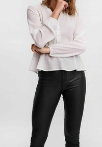 Weiße, strukturierte Bluse mit langen Ärmeln, Knopfverschluss und ausgestelltem Schnitt, kombiniert mit schwarzen, engen Hose aus einem glatten Material.