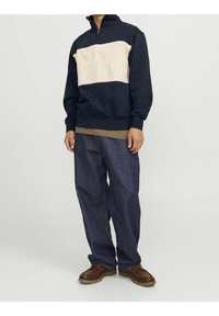 Hombre vestido con un sudadera azul marino y beige con el texto "Originals", jeans oscuros holgados, zapatos de cuero marrón, de pie contra un fondo liso.
