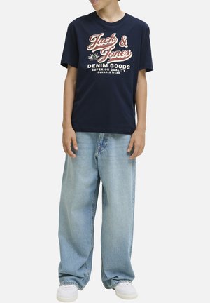 T-shirt en coton bleu marine avec un texte graphique blanc et rouge "Jack & Jones DENIM GOODS" et un jean en denim bleu clair décontracté. Baskets blanches.