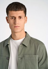 Groene lichtgewicht jas met knoopsluiting en een borstzak, gedragen over een eenvoudig wit T-shirt, op een effen achtergrond.