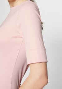 Primer plano de una persona que lleva una blusa ajustada de color rosa claro con mangas de longitud hasta el codo dobladas en los puños, contra un fondo liso.