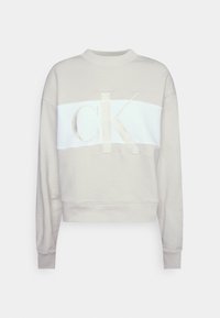 Sudadera corta color beige claro con una franja horizontal blanca y un gran logo "CK" bordado en el frente, mangas largas y puños acanalados.