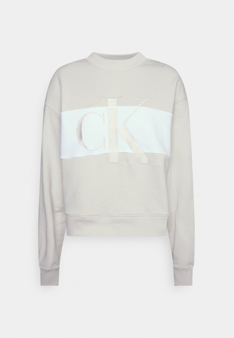 Sudadera corta color beige claro con una franja horizontal blanca y un gran logo "CK" bordado en el frente, mangas largas y puños acanalados.