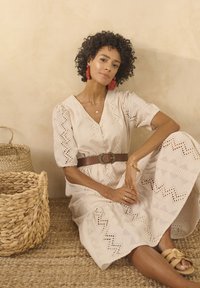 Vestito color crema con ricami intricati, chiusura a bottoni, maniche a sbuffo e motivi a zigzag. Cintura marrone con fibbia rotonda. Cesti intrecciati nelle vicinanze.