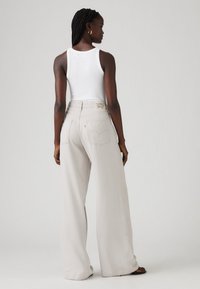 Lys grå vide bukser med høj talje, der har baglommer og et synligt mærke. Modellen har en hvid tanktop på.