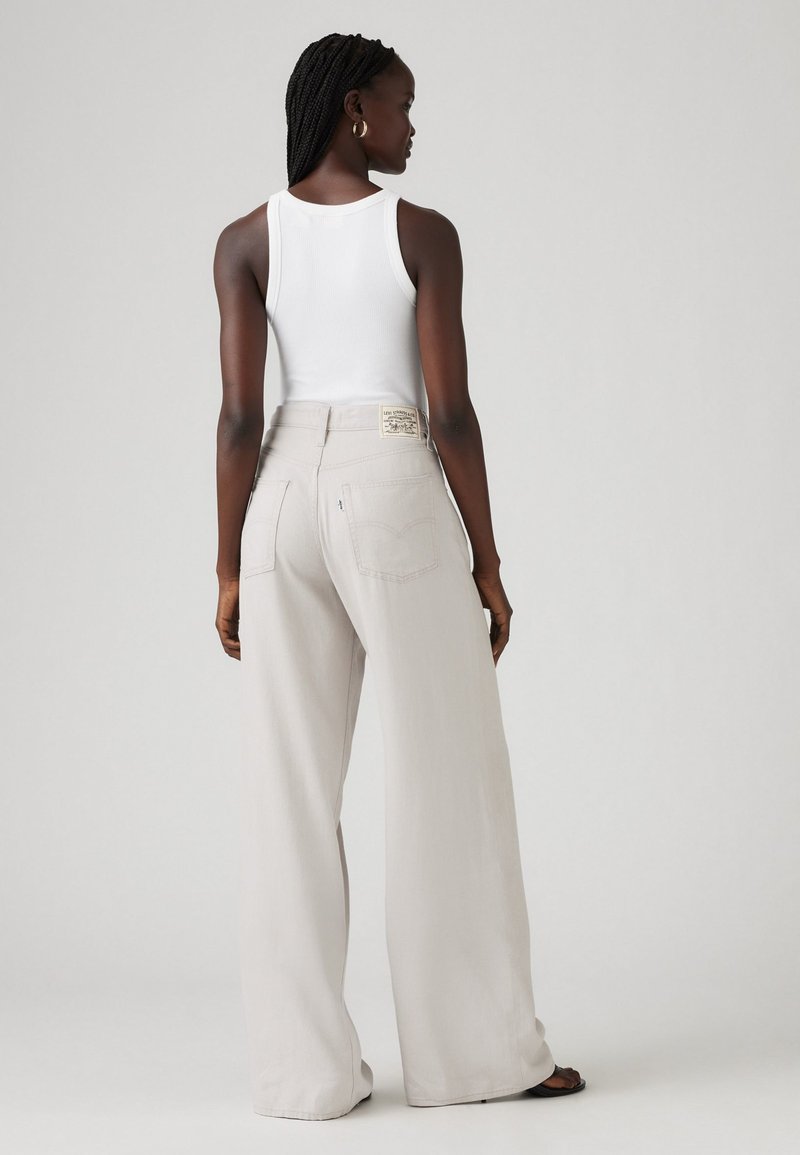 Pantaloni larghi grigio chiaro con vita alta, dotati di tasche posteriori e di un'etichetta del marchio visibile. Il modello indossa una canotta bianca.
