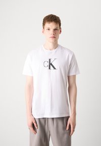 Balta medvilninė marškinėliai su ryškiu juodu "cK" logotipu krūtinėje. Trumpomis rankovėmis ir apykakle. Lygus paviršius.