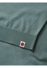 Suéter verde claro con dobladillo acanalado y una pequeña etiqueta blanca con un punto rojo. Textura suave con un diseño clásico y sencillo.