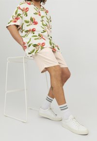 T-shirt à manches courtes avec un motif de pêches et de feuilles, associé à un short rose clair et des baskets blanches avec des chaussettes rayées grises, assis sur un tabouret.
