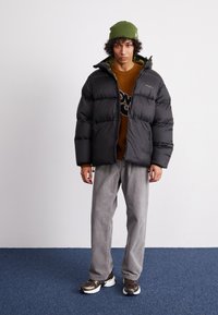 Schwarze Daunenjacke mit Reißverschluss vorne und zwei Seitentaschen, über einem braunen Pullover und grauen Cordhosen getragen. Grüne Strickmütze auf dem Kopf.