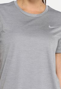 Camiseta deportiva gris con cuello redondo y mangas cortas. Presenta un pequeño logo reflectante de Nike en el pecho. Textura suave.