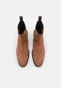 Bottines en daim marron au design élégant, avec des panneaux latéraux élastiques et une semelle en caoutchouc noire. Dotées d'une languette de tirage avec des accents rouges.