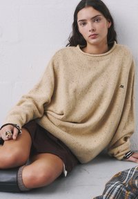 Pulover bej tricotat, cu o croială lejeră și guler cu zimți, având o textură pătata și un logo mic. Asortat cu o fustă închisă și șosete gri.