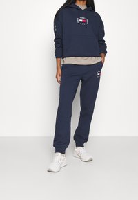 Marinblå hoodie med framficka och logotyp, i kombination med matchande sweatpants. Vita sneakers fulländar den avslappnade outfiten. Material av bomullsblandning.