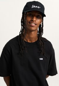 Schwarzes Baumwoll-T-Shirt mit weißem "VANS"-Logo auf der Brust, kombiniert mit einer passenden schwarzen Cap, die dasselbe Logo in weißer Schreibschrift zeigt.